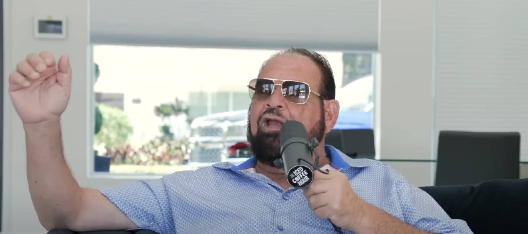 Man in sunglasses sits stretched out on couch, holding microphone and gesturing broadly with his other arm.