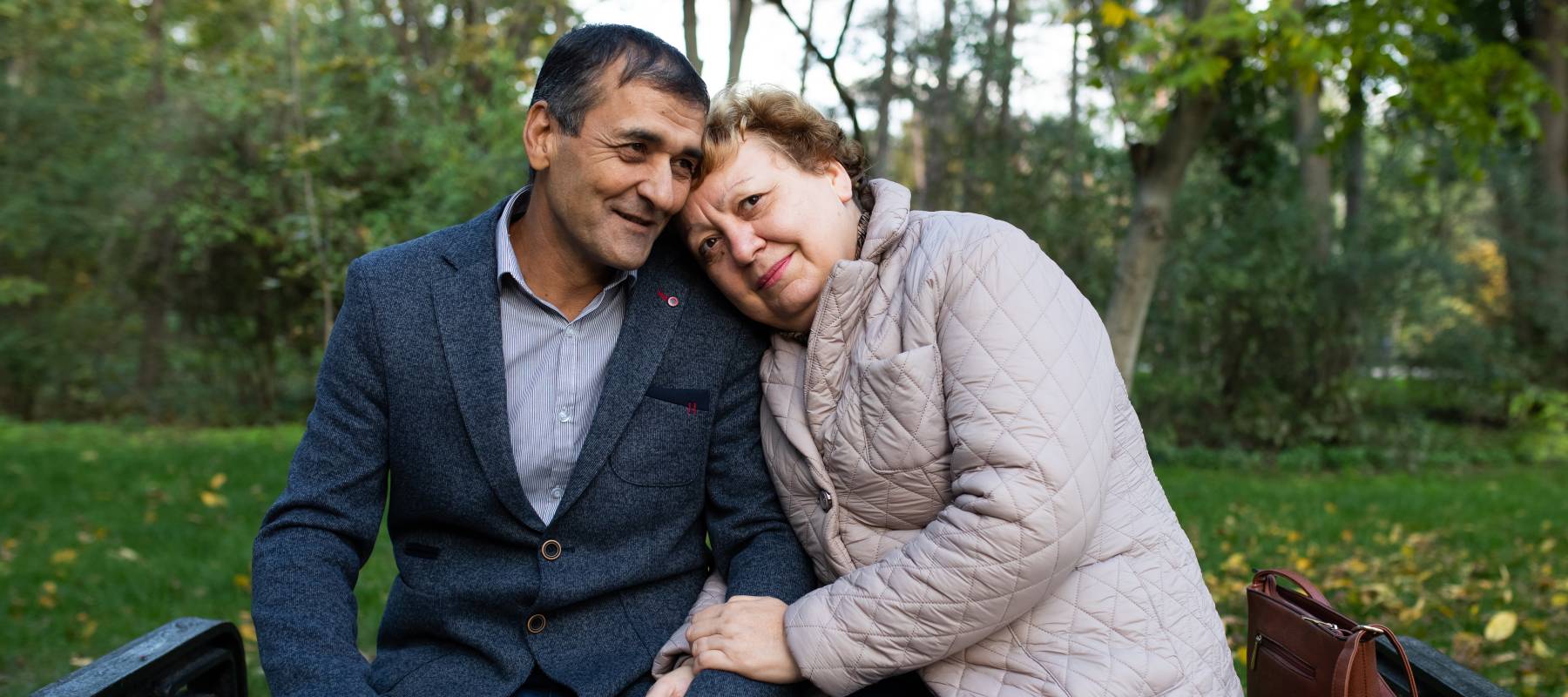 Older couple sitting on a park bench, woman's head on man's shoulders and looking at the camera directly.