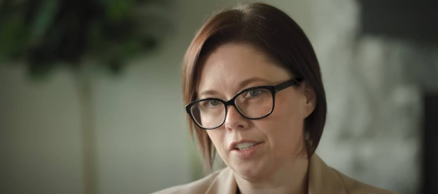 Woman in glasses speaking to the camera, looking serious.