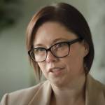 Woman in glasses speaking to the camera, looking serious.