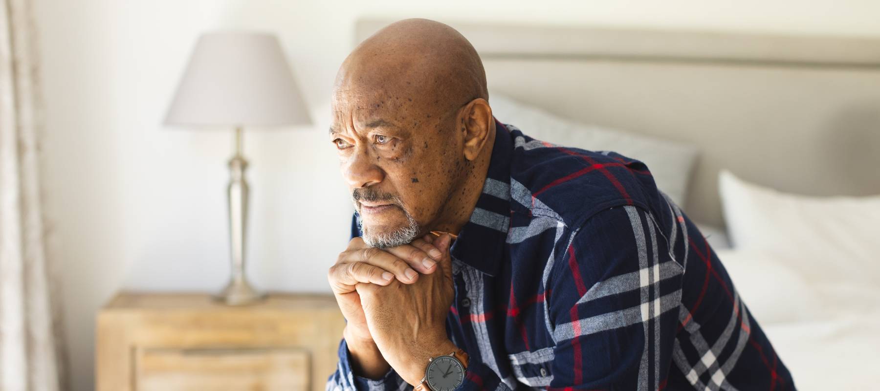 pensive African-American senior man sitting on bed in sunny bedroom