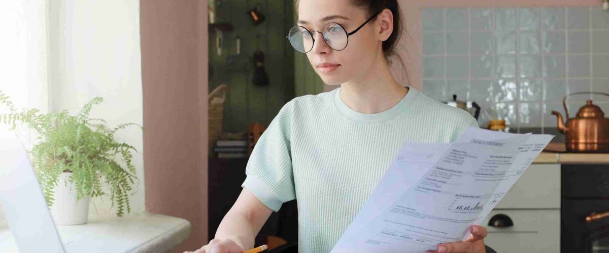 Young woman managing domestic budget, sitting at kitchen table with open laptop, documents and calculator, using touchpad, making notes with pencil