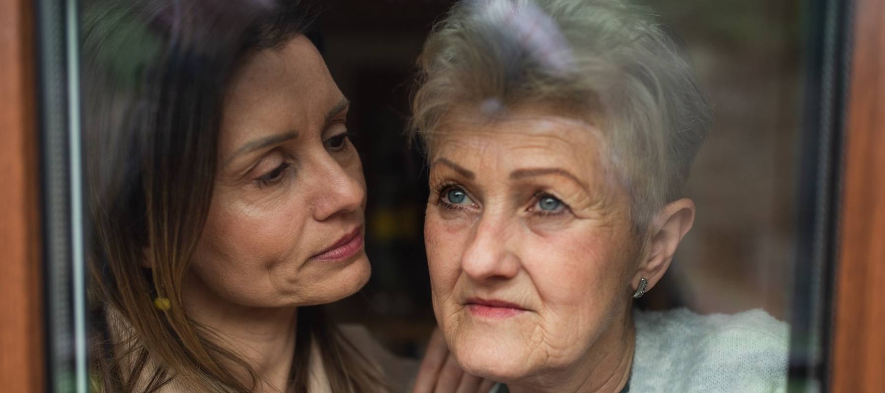 Close-up portrait of sad senior mother with adult daughter indoors at home, hugging