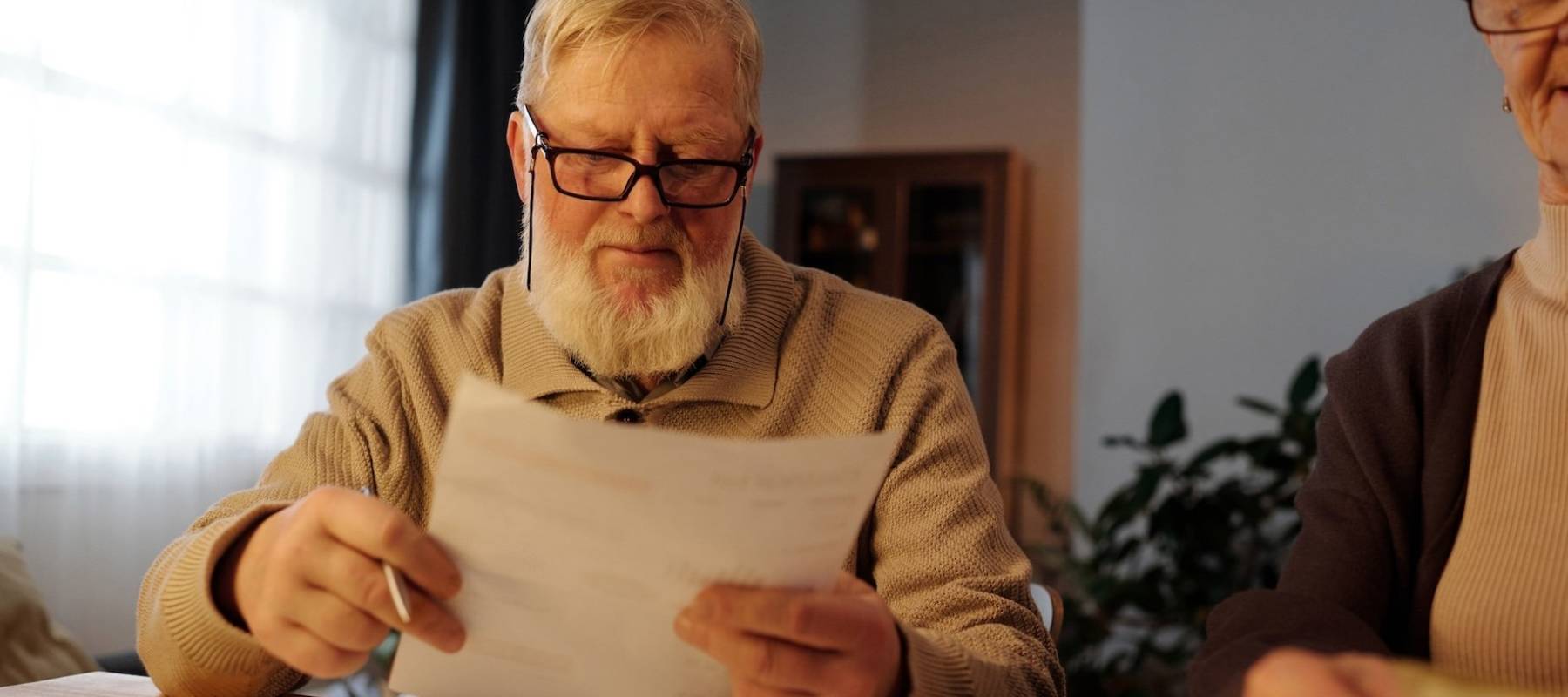 Serious mature man with white beard holding financial document with housing payment sums and looking through information