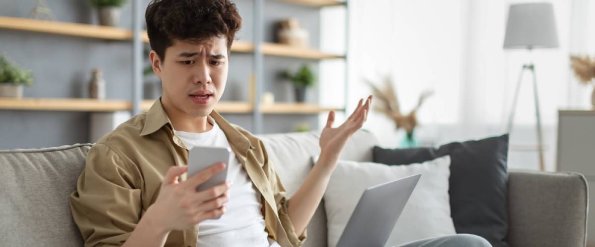 confused Asian man looking at phone screen, sitting on couch with computer on lap