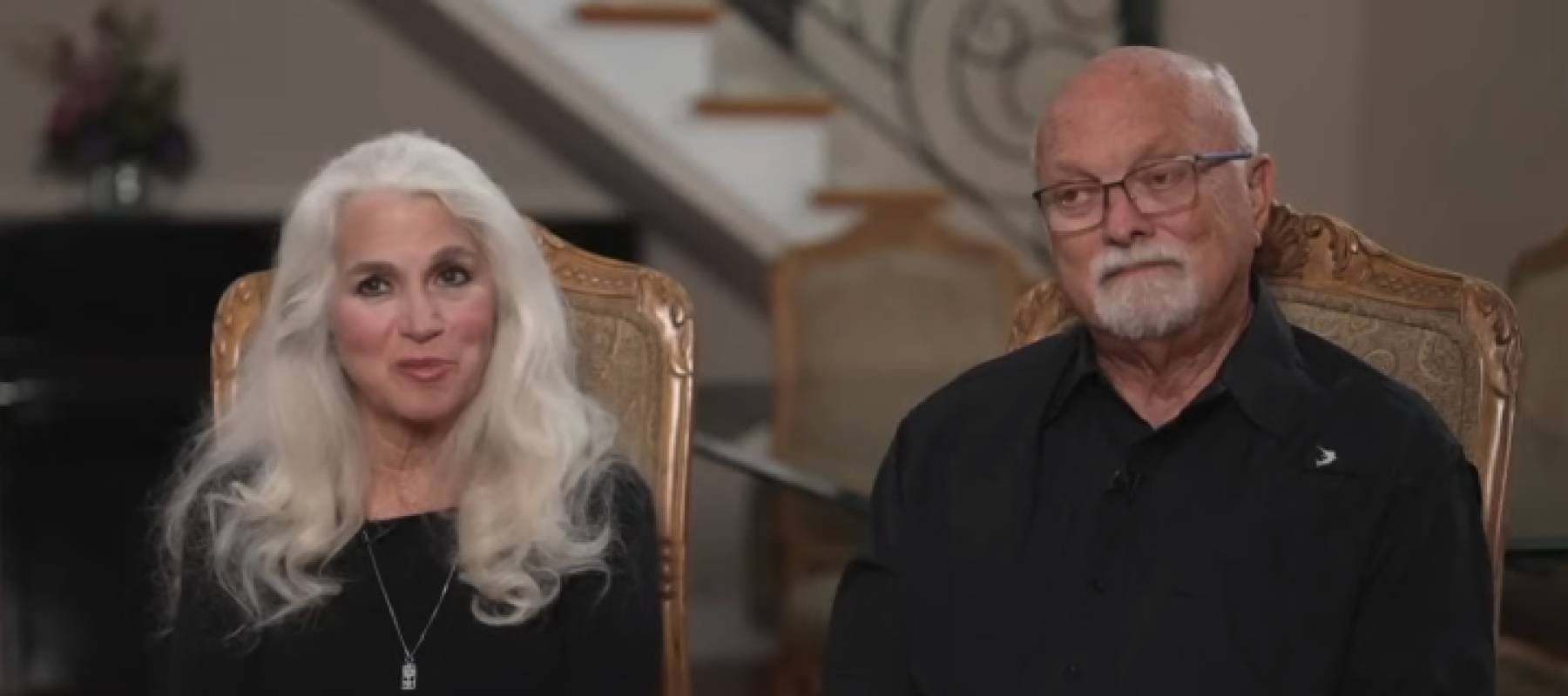 Couple sit on chairs in dining room, speaking directly to camera, looking concerned.