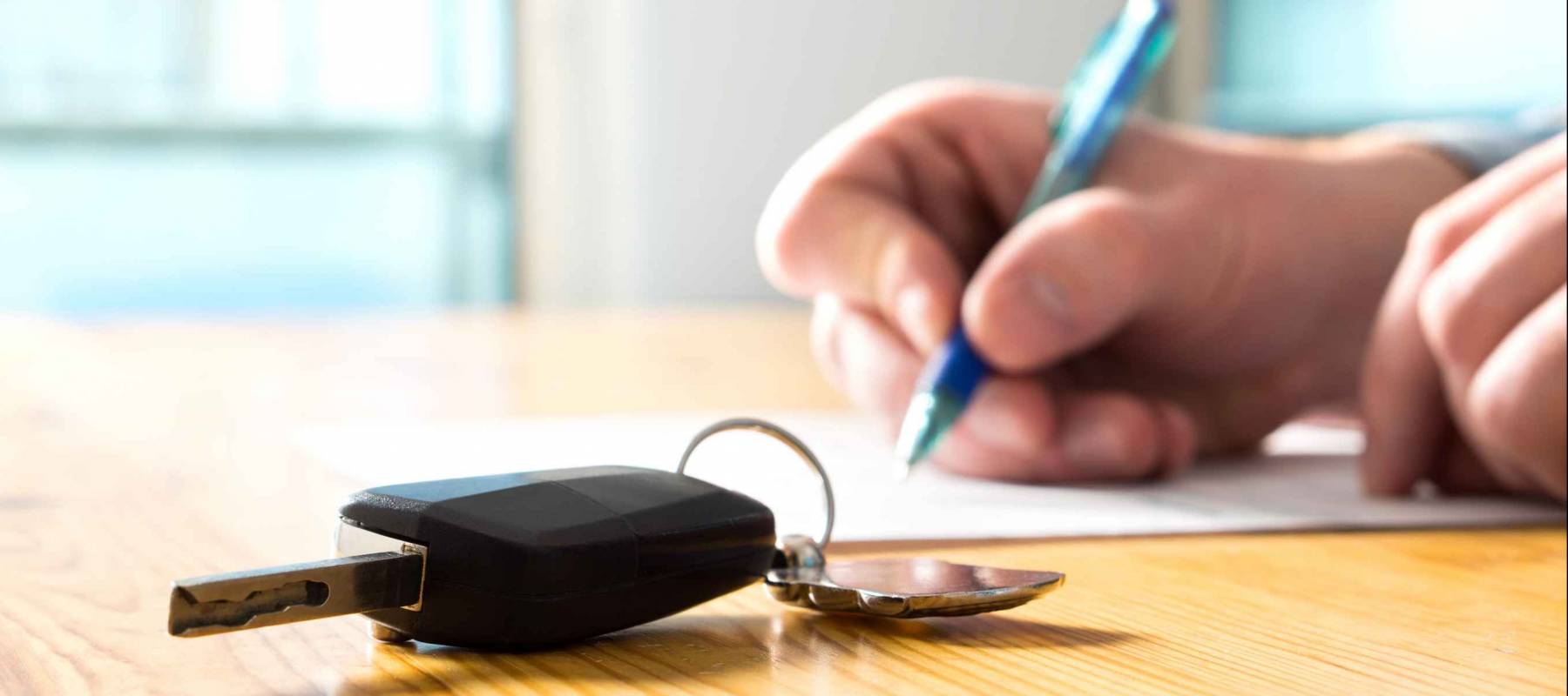 A hand signing a document next to a car key laying on a table