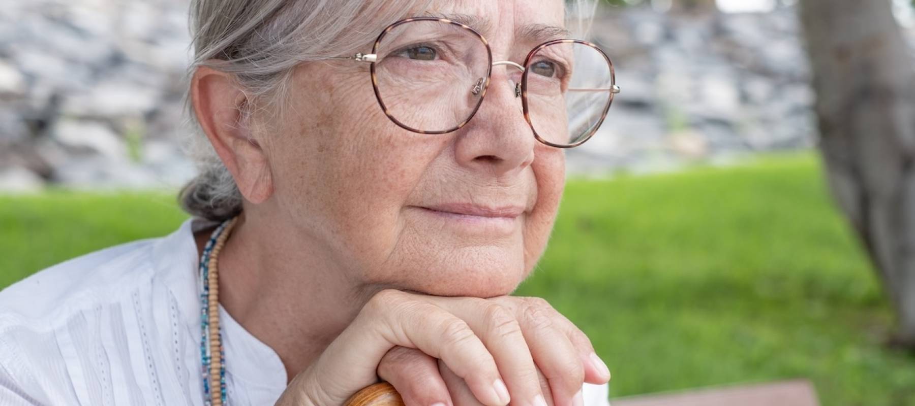 Portrait of a thoughtful elderly woman of 70 years old sitting on a bench in the park leaning on a stick due to walking disability