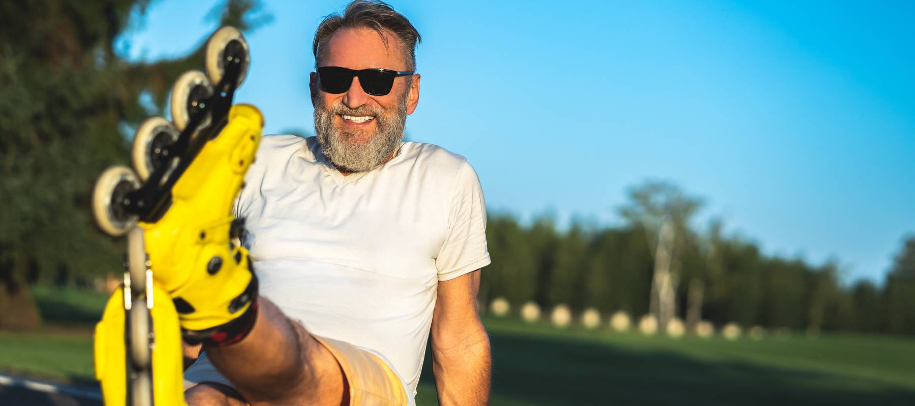 Happy older man sits on pavement, leaning back on his arms and smiling with legs crossed, rollerblades on his feet.