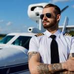 Cool young pilot in sunglasses with tattoos on his hands posing at private air plane on runway. Good summer weather, handsome male model