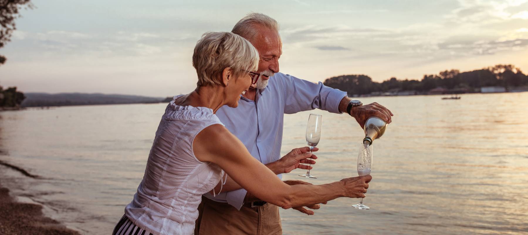 Senior couple stand on beach, pouring glasses of champagne and smiling.
