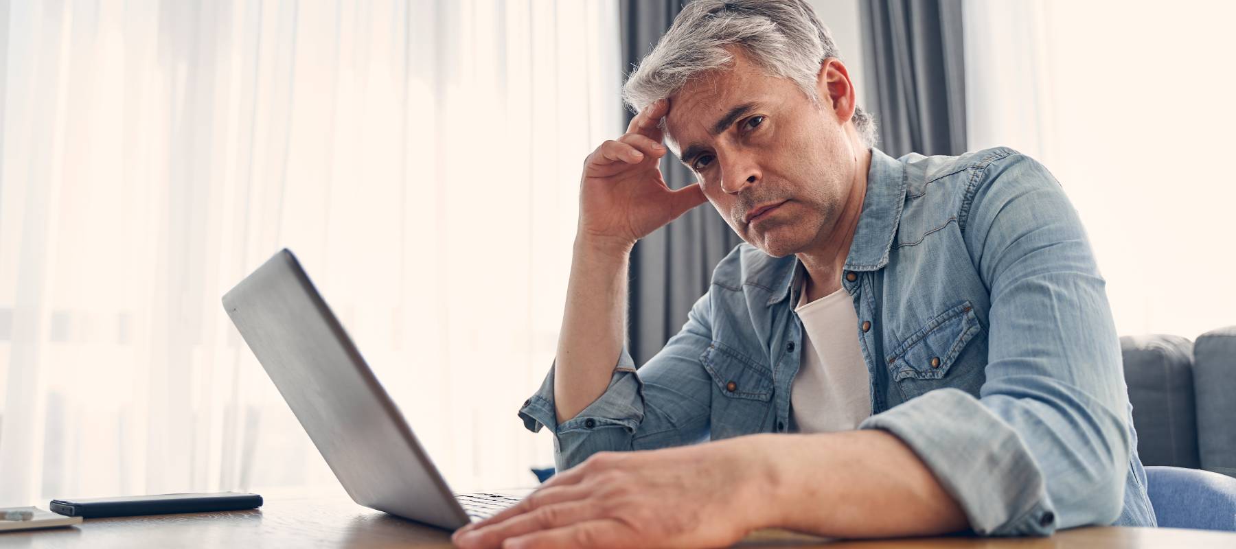 Older man sits at kitchen table with head in hand, looking directly at camera with laptop open in front of him.