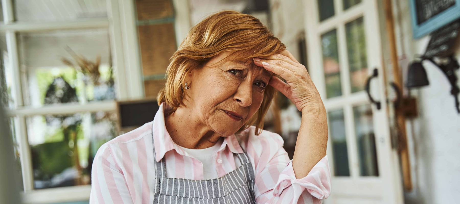 Older woman in an apron holds her hands to her face and looks at camera with worry, disappointment in her eyes.