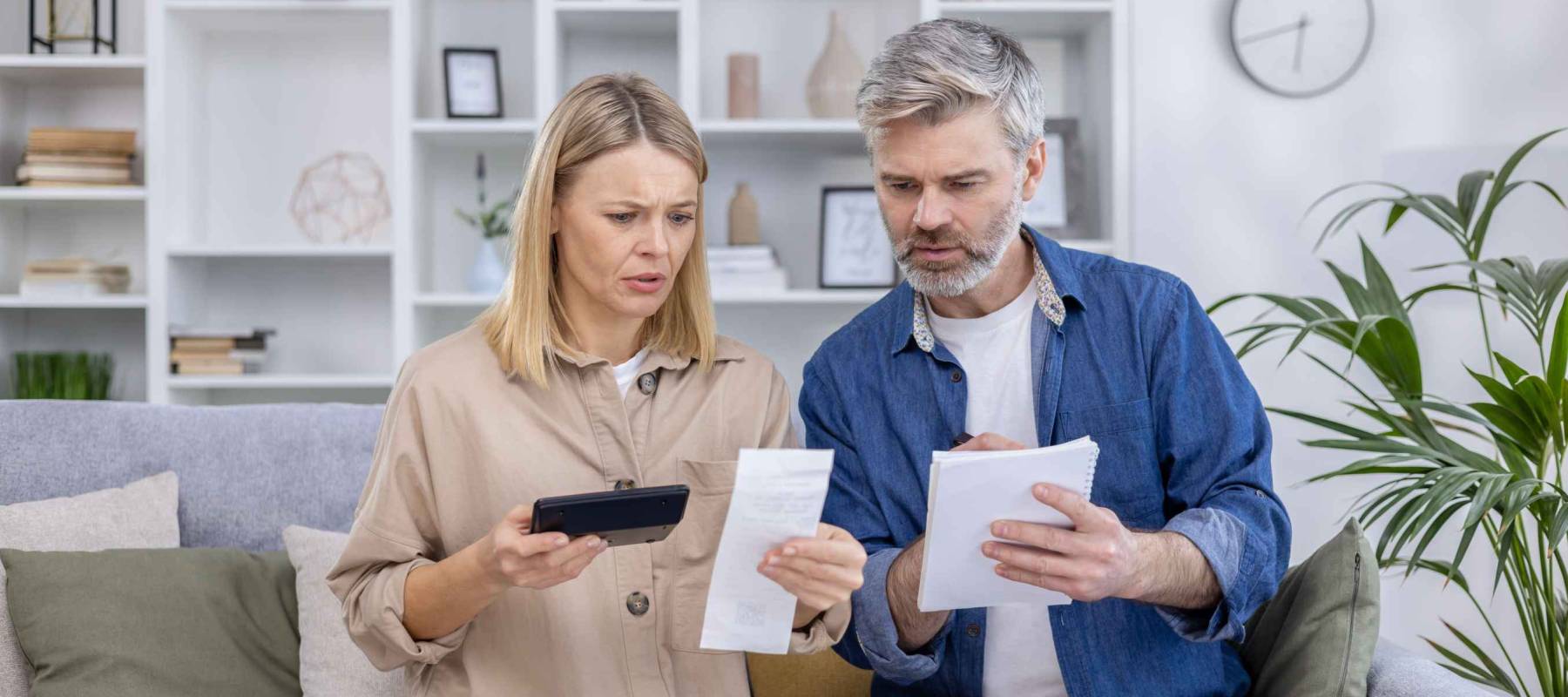 Worried mature couple reviewing bills together at home, calculating finances with a smartphone and notepad. Concept of financial planning and family budgeting.