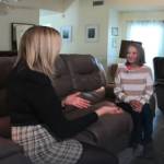 Two women sit facing each other on a couch in living room, talking.