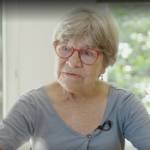 Older woman in glasses sits at table and talks to reporter.