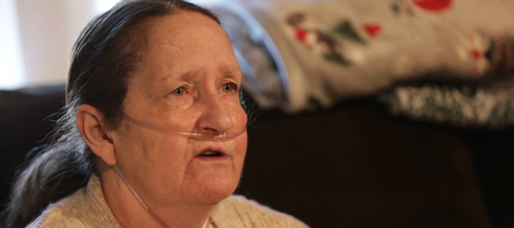 Older woman seen sitting on couch, talking to camera, looking upset. Wearing an oxygen mask.