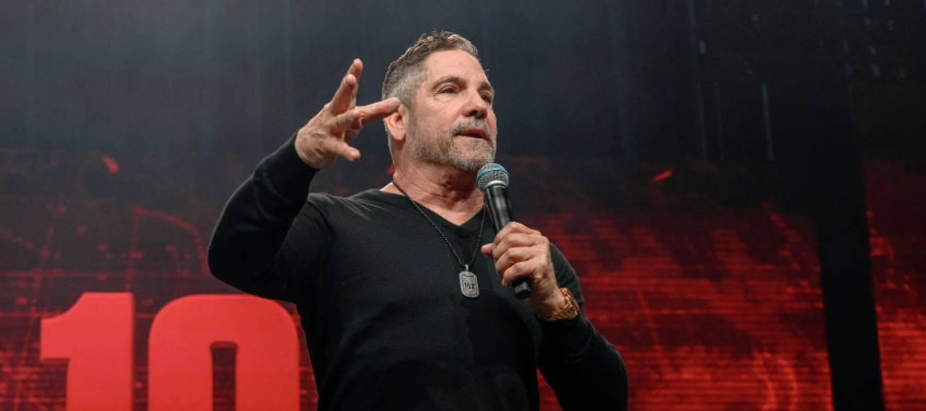 Grant Cardone seen on stage speaking into a microphone, holding out his other hand with palm facing the ground.