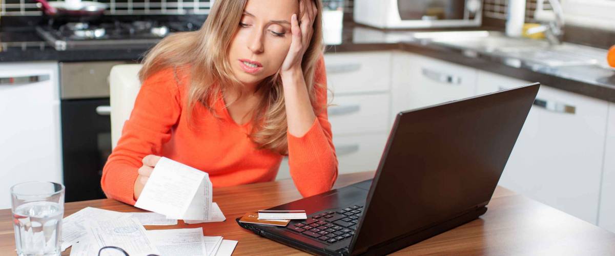 stressed over bills. portrait of a young caucasian woman using a laptop computer sitting at her kitchen holding utility bills and bank statements being thoughtful and worried. Home interior.