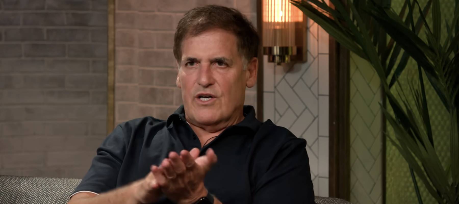 Mark Cuban speaks on podcast, sitting on couch, making gesture with hands.