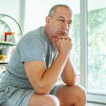 Sad middle-aged man at home sitting on bed in bedroom.