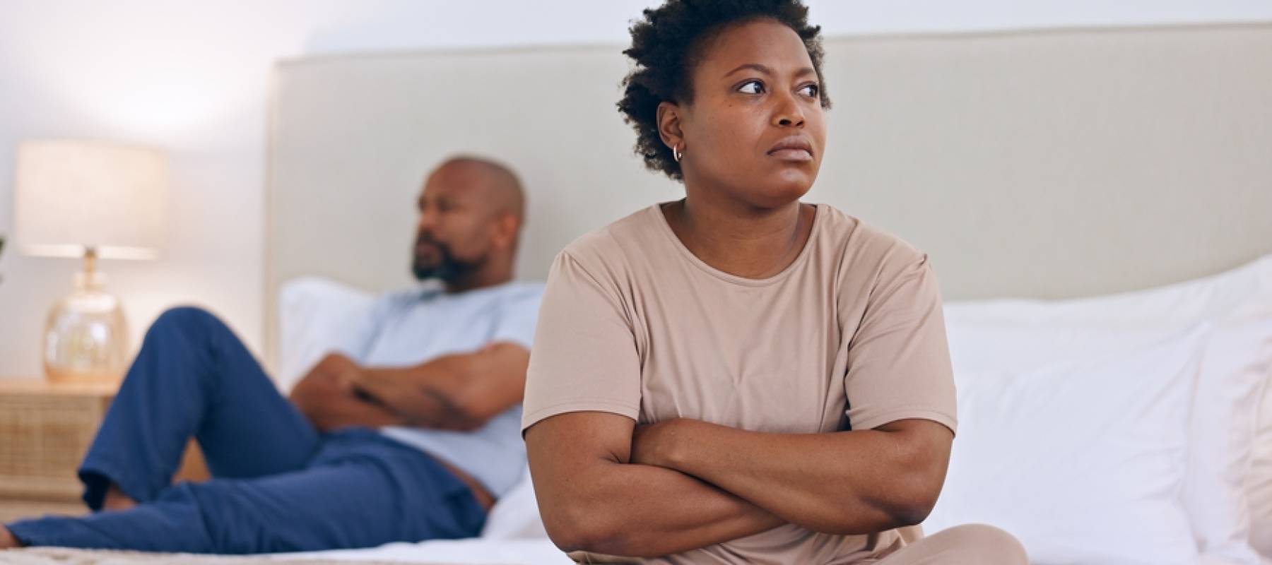 An unhappy couple sits on a bed, not speaking.