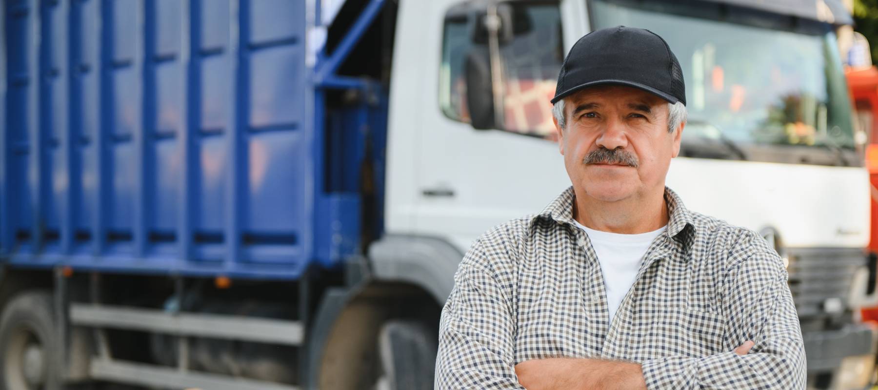 A male garbage truck driver is standing near the cab with arms crossed, looking serious.