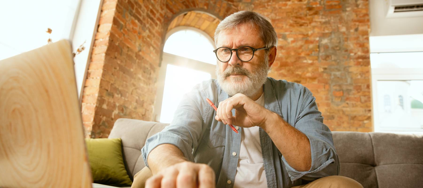 Bespectacled senior with grey hair and beard looking pensively at a laptop, holding a pen.