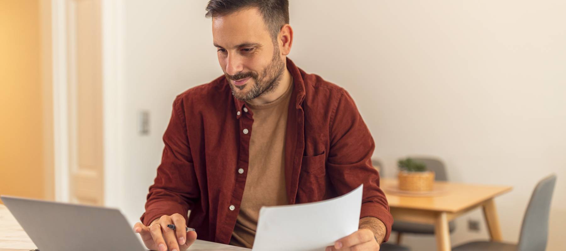 A 40ish-year old man with dark hair and facial hair looking at his laptop with a paper in his hand.