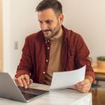 A 40ish-year old man with dark hair and facial hair looking at his laptop with a paper in his hand.