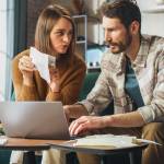 Man and woman sitting in front of a computer and talking, as woman holds and points to a piece of paper.