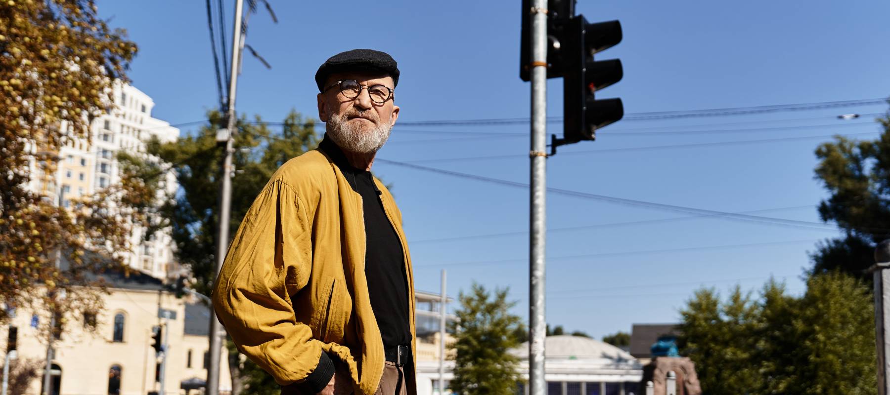 A stylish senior man walks through an urban environment, looking directly at the camera and posing.