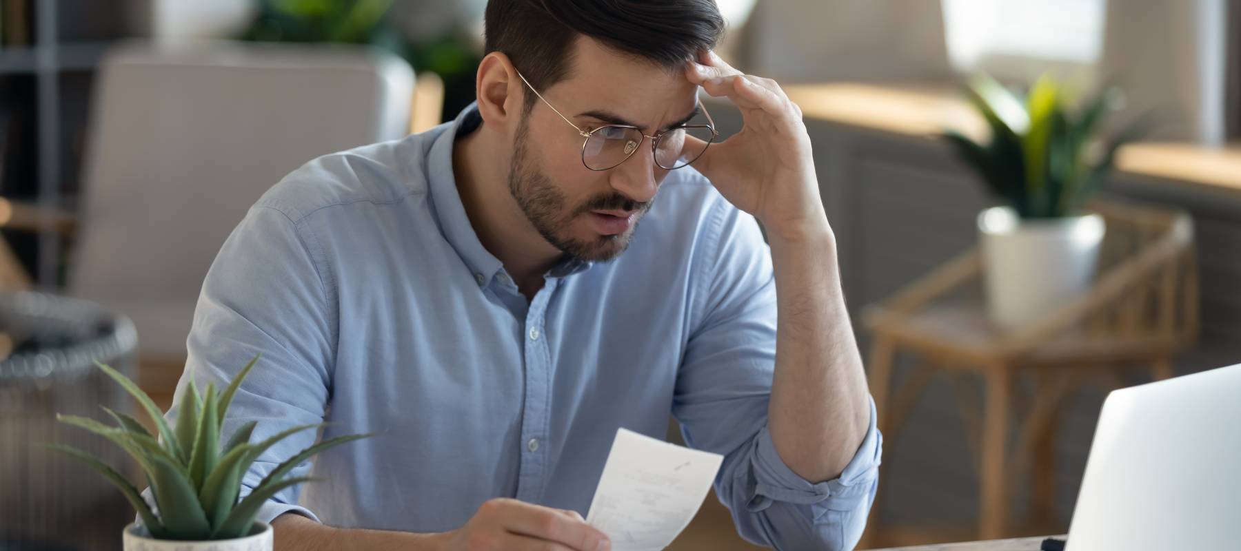 A 40ish man with glasses holding a receipt and staring at his laptop in concern.
