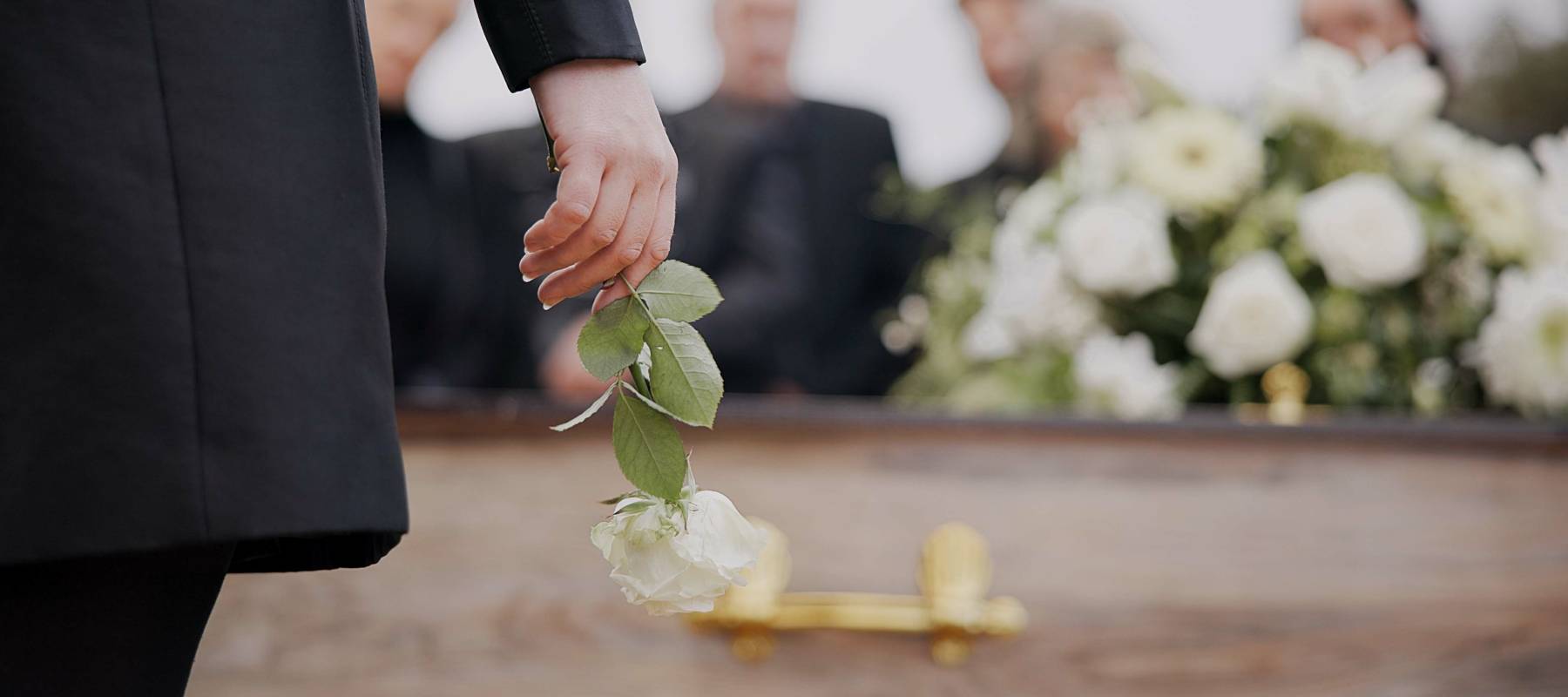 Hand holding a white rose in front of a casket with another flower arrangement on it.