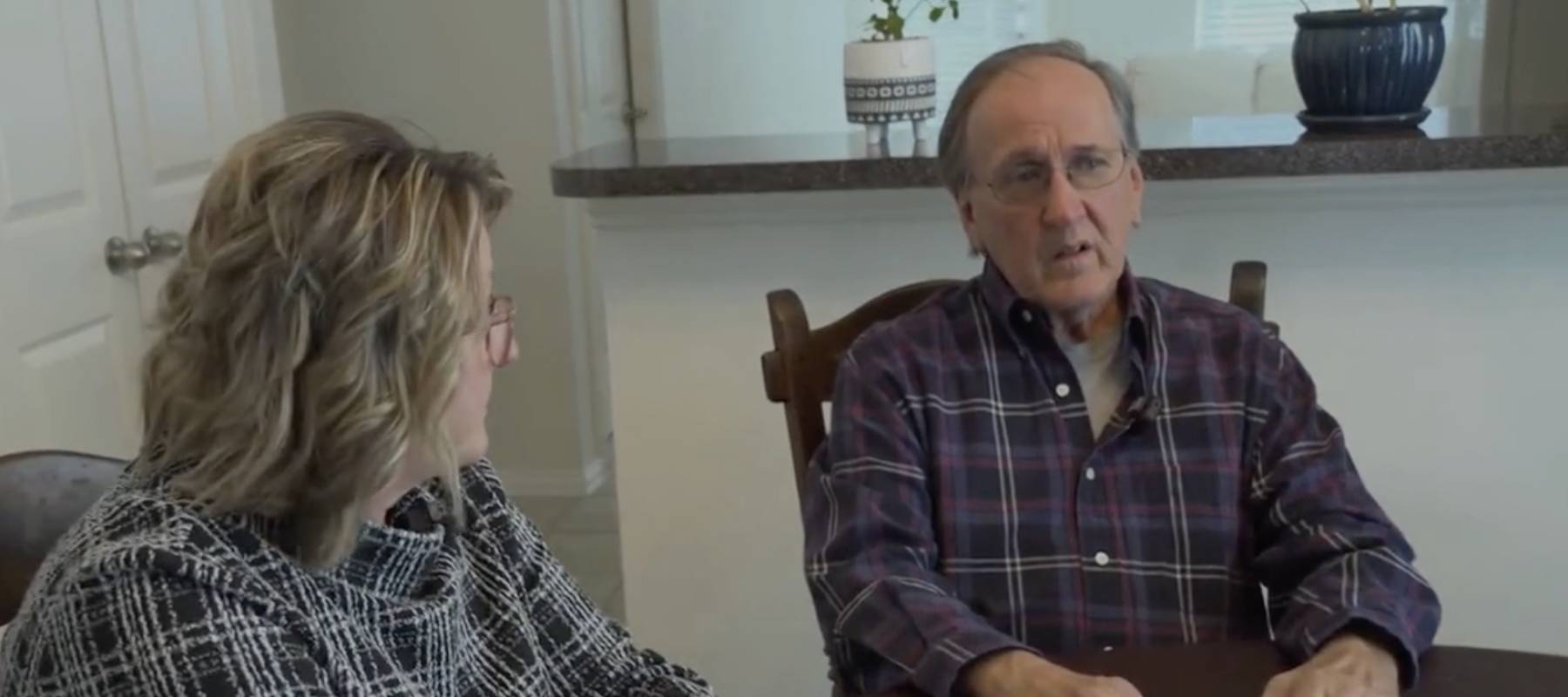 Man and woman sit at kitchen table, talking. Man looks distressed.