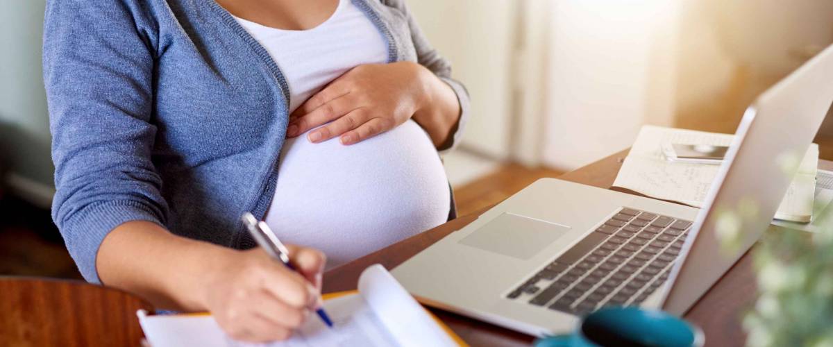 Closeup of a pregnant woman writing notes and using a laptop while working on maternity leave at her dining room table at home