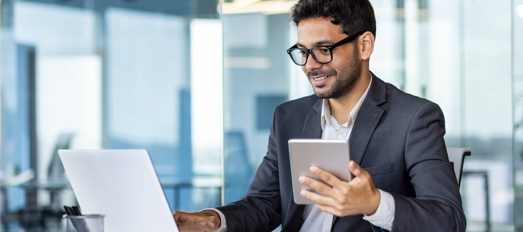 Mid-thirties man in an office environment with a partial smile looking at his laptop while holding a tablet.