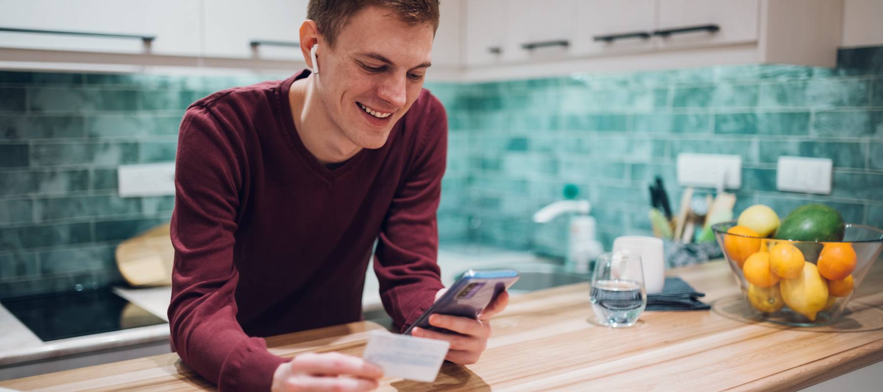 Young man in kitchen looking at finances on smart phone holding credit card