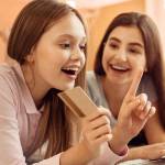 Incurable shopaholic. Excited pretty teenage girl lying on the bed next to her elder sister and choosing an item in an online shop while holding a golden credit card