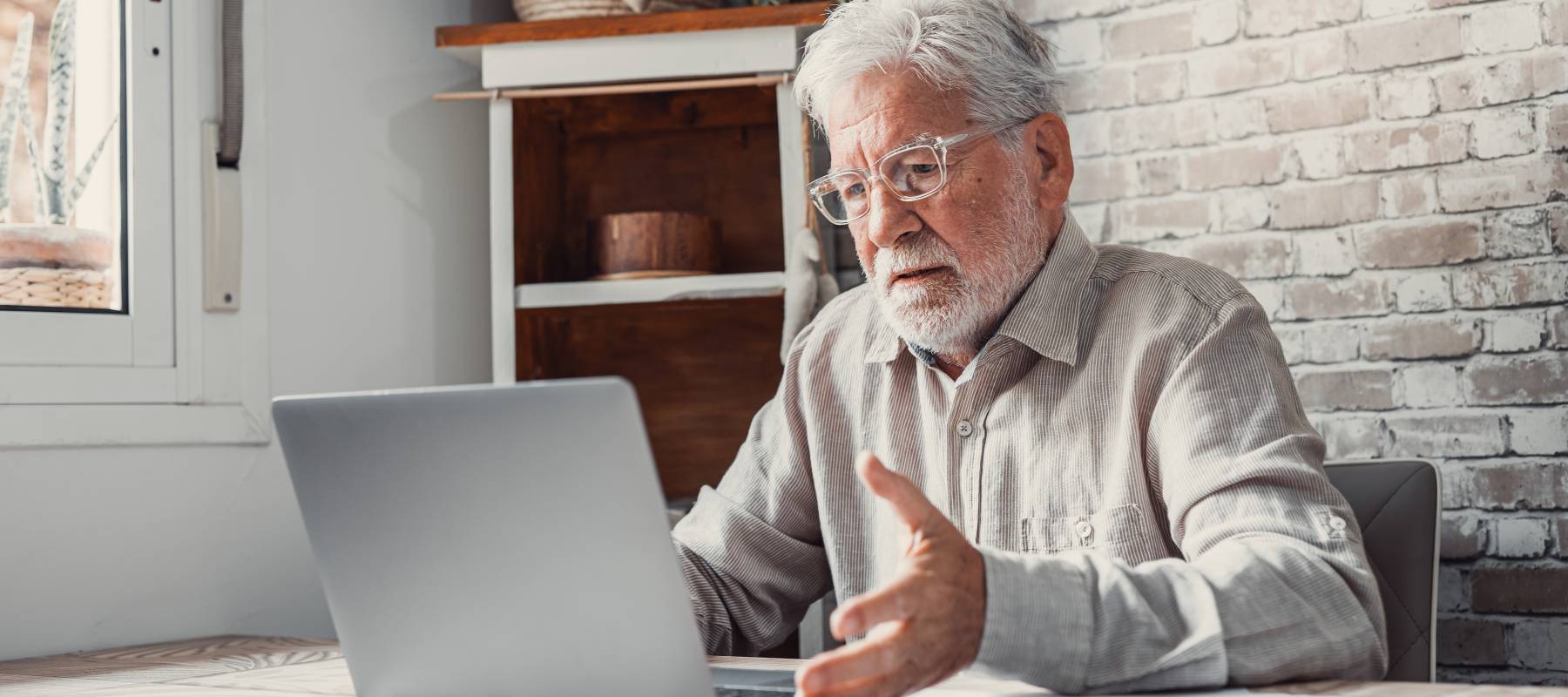 Elderly bespectacled gentleman with white hair and beard sitting in front of his laptop in dismay.