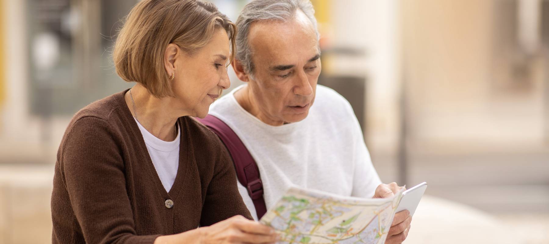 Retirement Tourism Concept. Mature European Tourists Couple Looking At Touristic Paper Map Choosing Travel Destination For Vacation Sitting Outside. Spouses Planning Journey Abroad Together