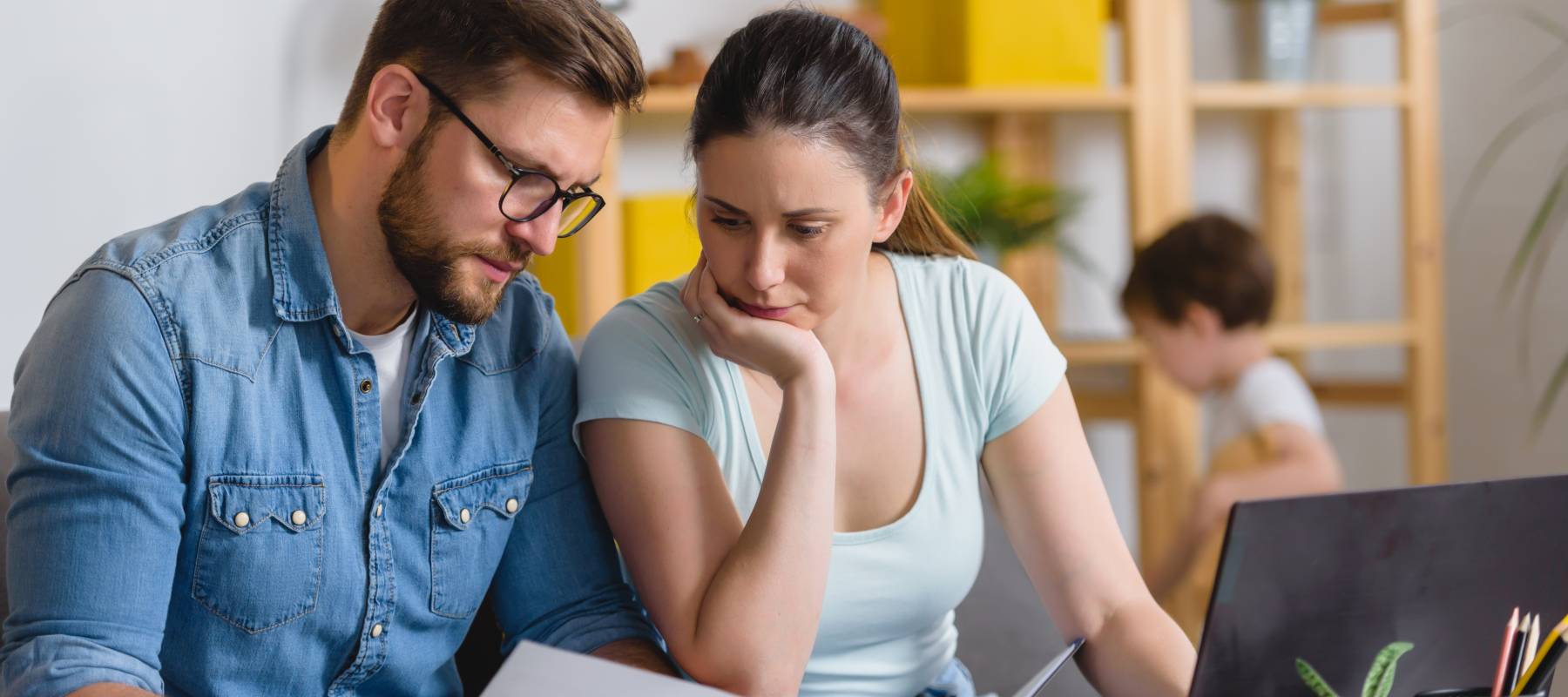 Worried mid-30s couple reviewing their finances in front of a laptop, with a preschooler child playing in the background.