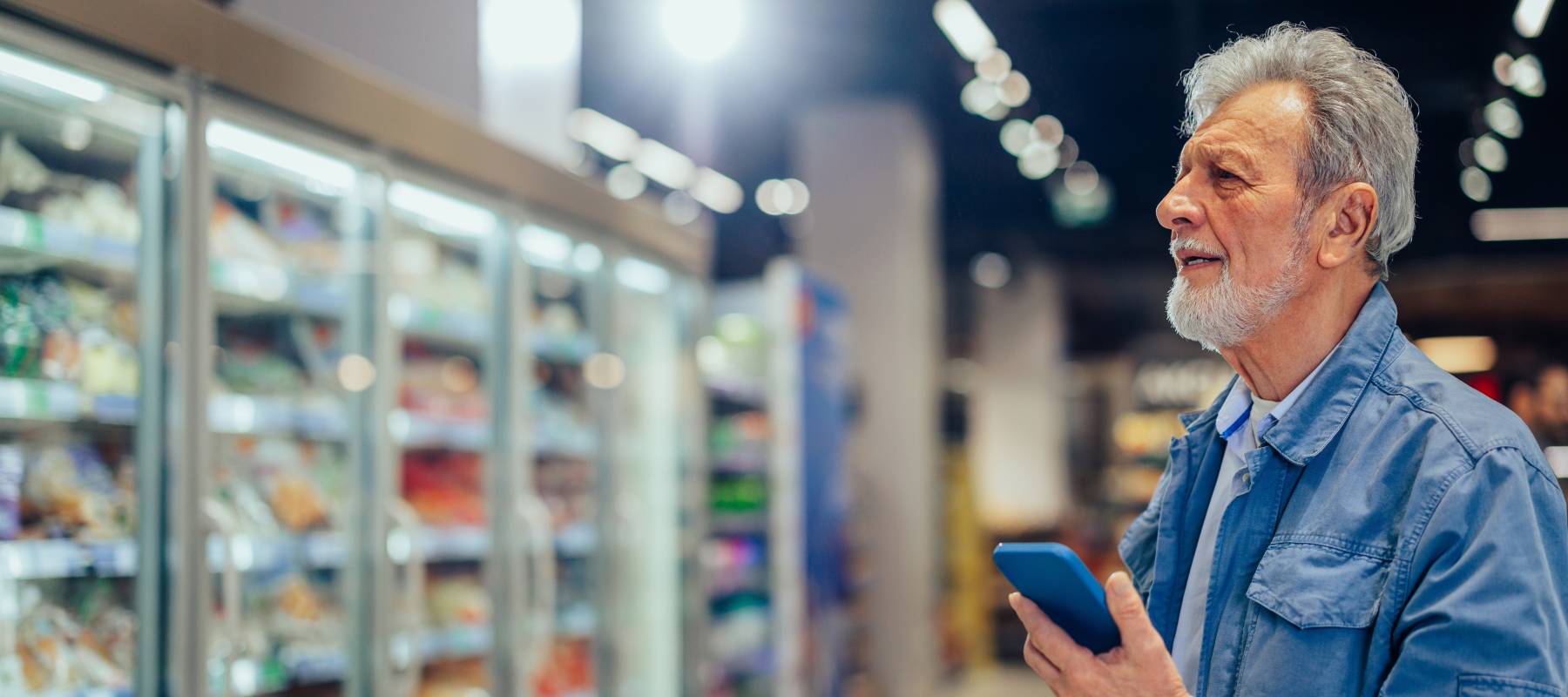 Gray-haired man pushing shopping trolley in market and checking coupons on his smartphone.
