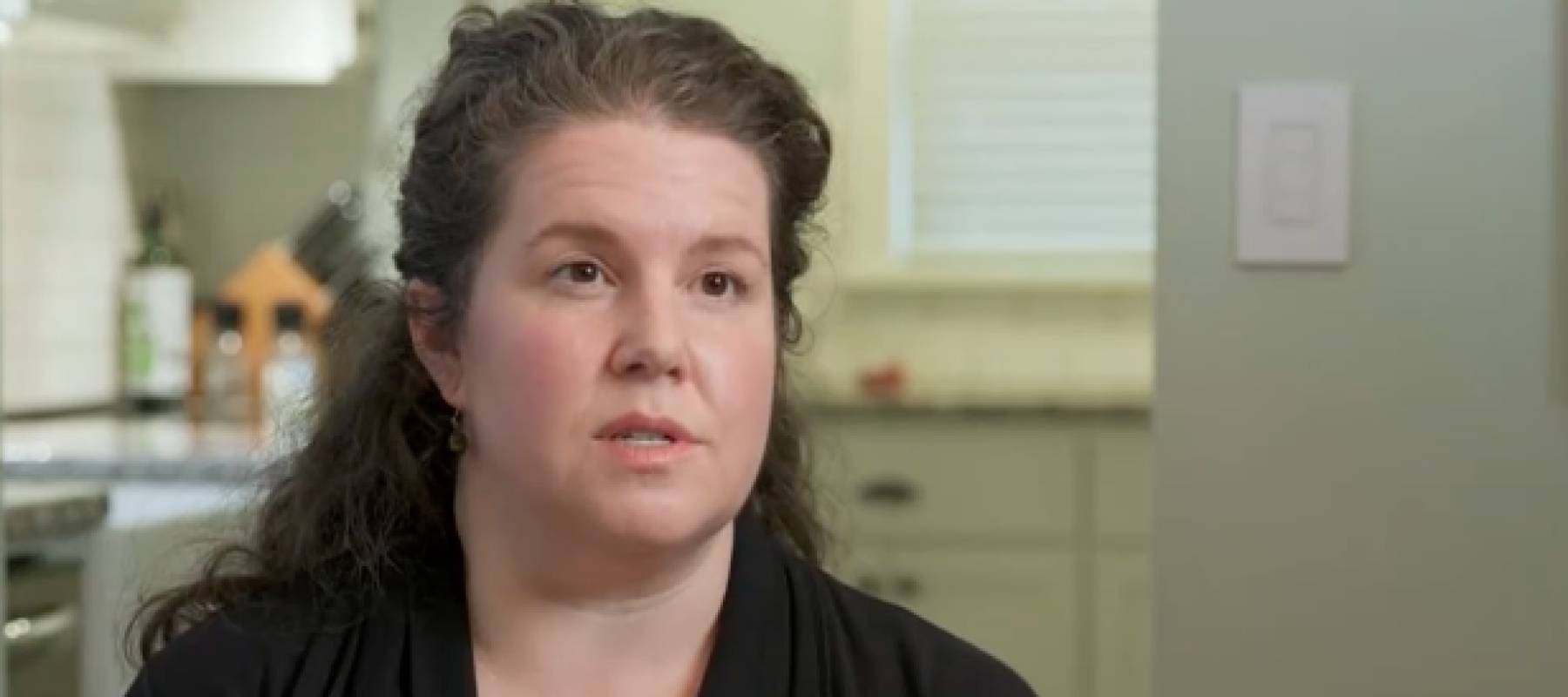 Woman sits in front of her kitchen, speaking on camera.