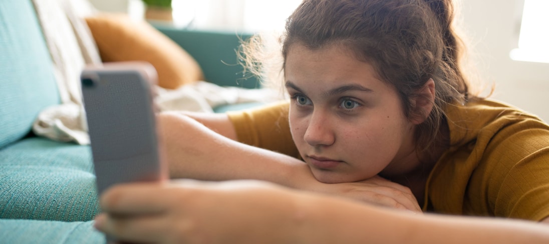 A young girl lays on a couch, holding and staring intensely into a phone screen.