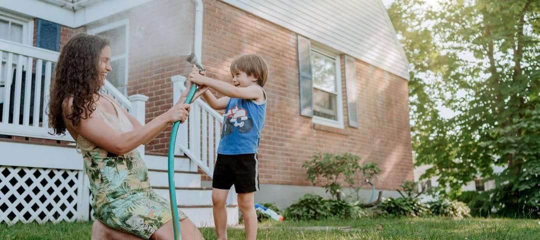 Woman with curly hair and young boy play with a hose outside an older home