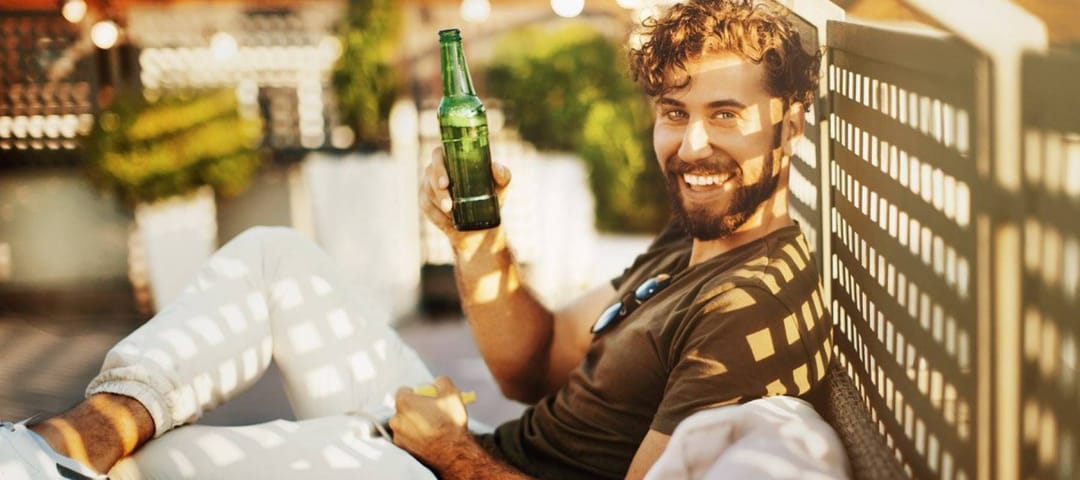 Smiling hipster with beard and curly hair holds up a bottle of beer