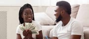Planning family budget. Happy afro couple sitting on floor at home with dollar cash and notepad, deciding together how to spend money