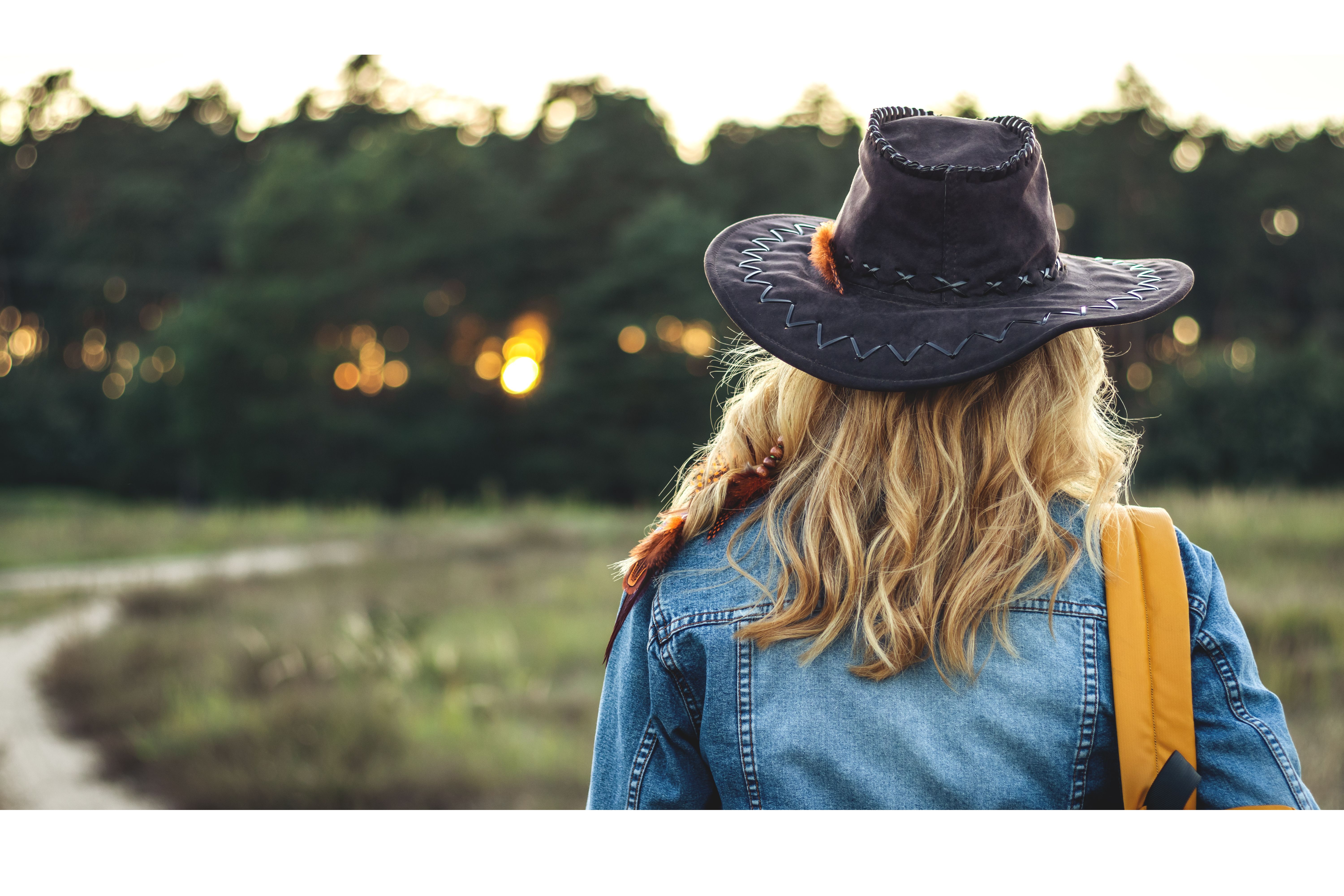 Woman tourist with hat and backpack hiking on footpath in nature during sunset. Rear view of blond hair female tourist wearing denim jacket is walking outdoors.