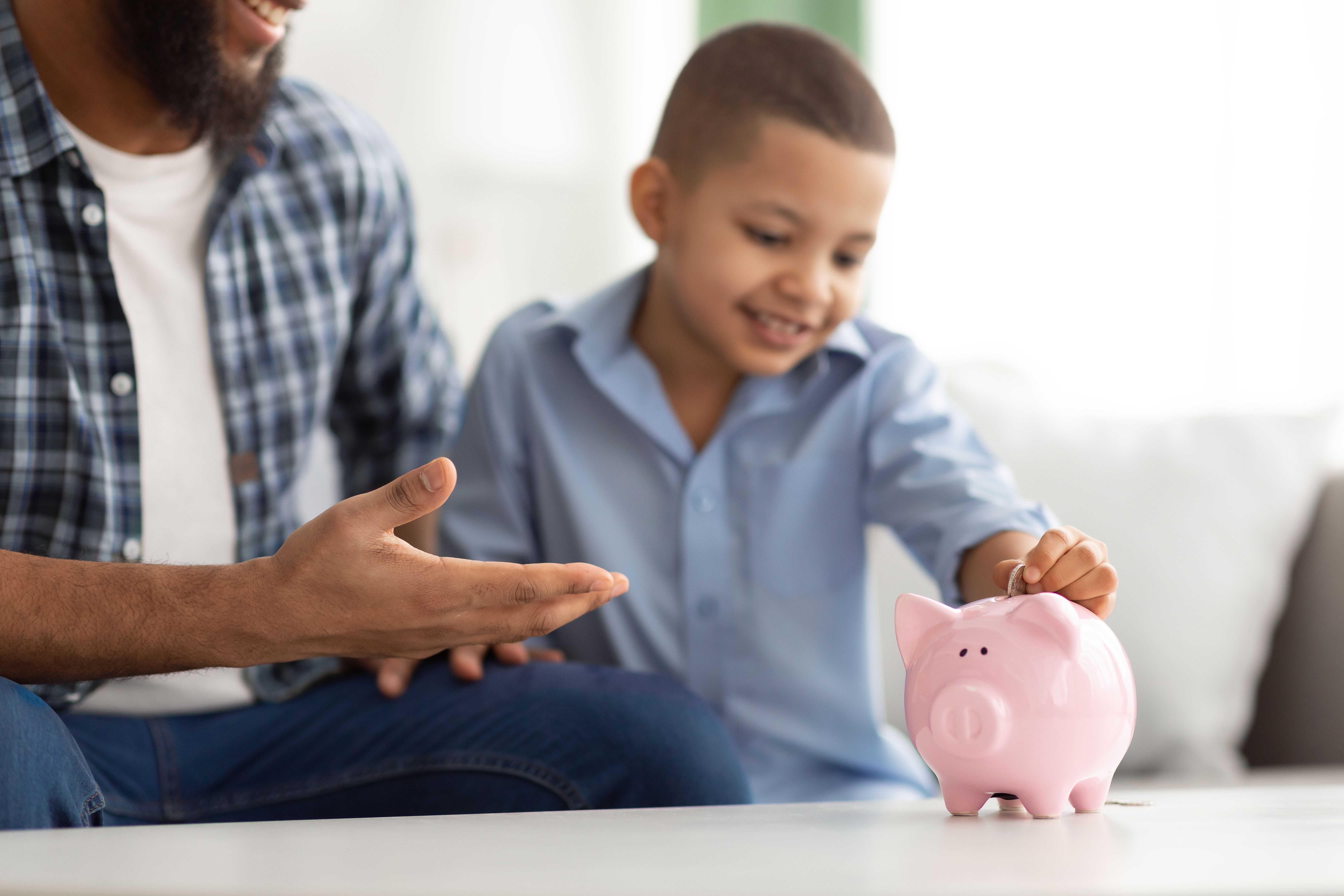 Personal Savings. Black Dad And Son Putting Coin In Piggybank Sitting At Home. Young Father Teaching His Child Financial Literacy And Budget Planning. Selective Focus On Piggy Bank, Cropped